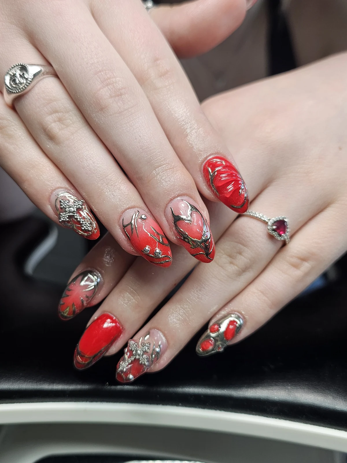 Red almond nails with nude bases, metallic silver linework, cross charms and raised red accents.