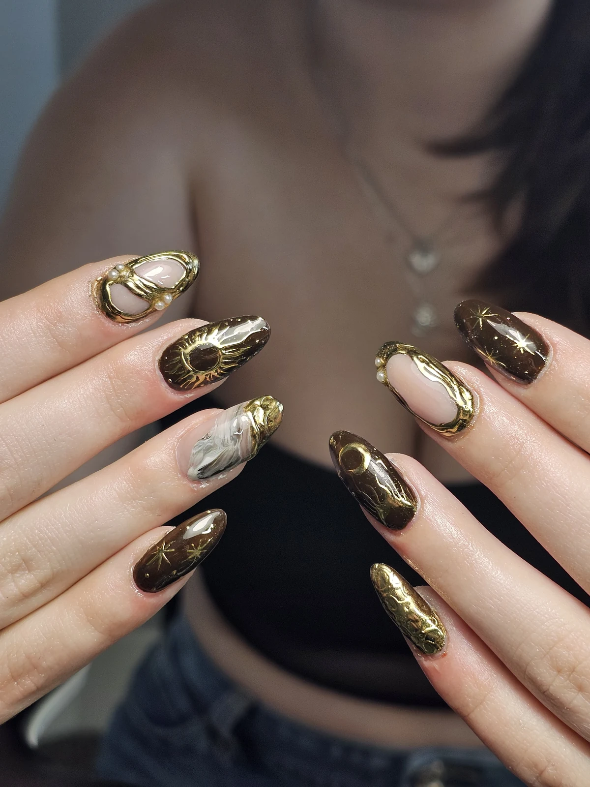 Brown and nude almond nails with gold sun, moon and star details, marbling and pearl accents.