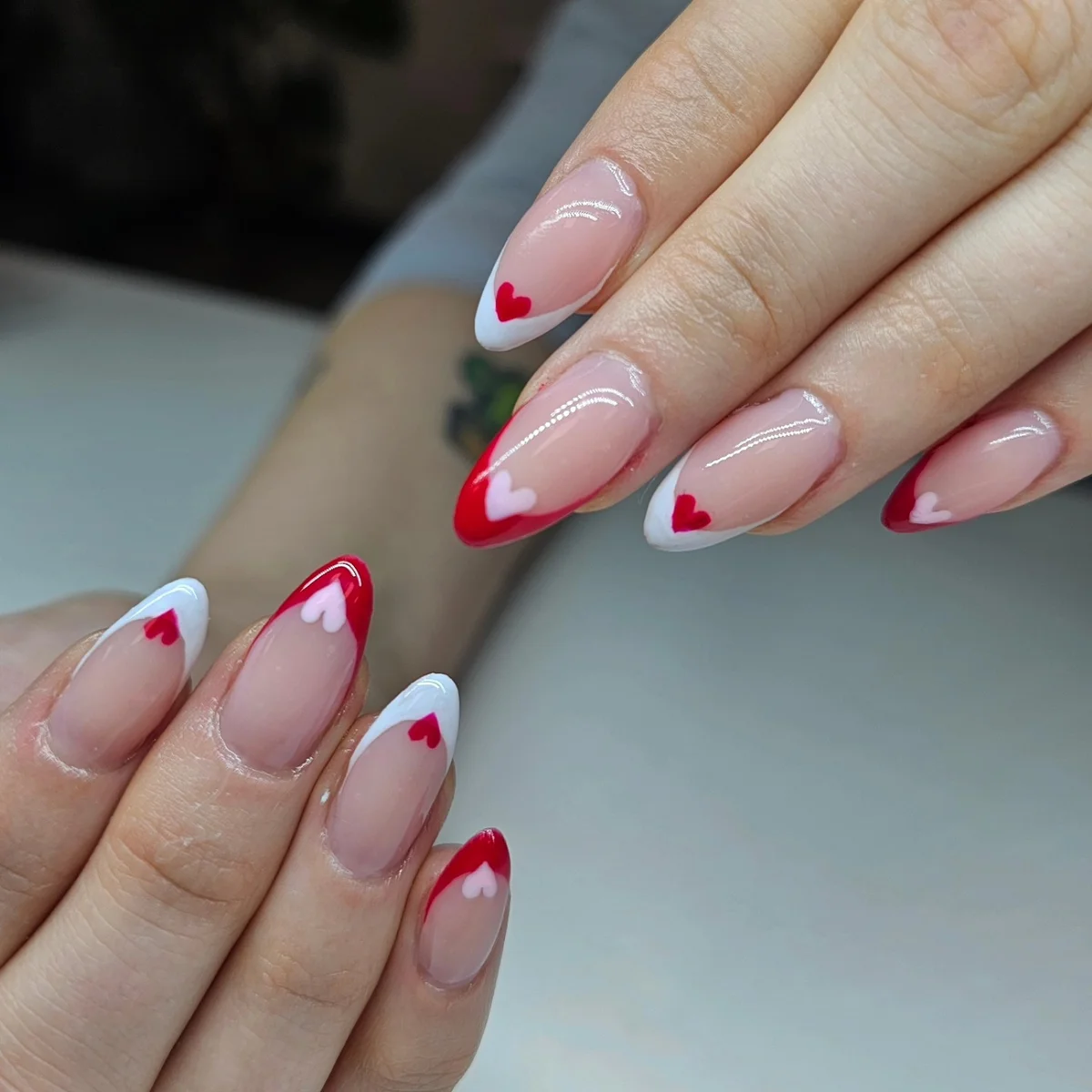 Nude almond nails with red and white French tips and small red hearts.