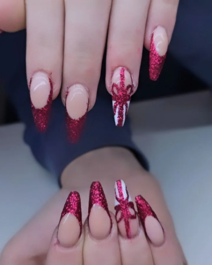 Long nude nails with red glitter French tips and red bow accents.